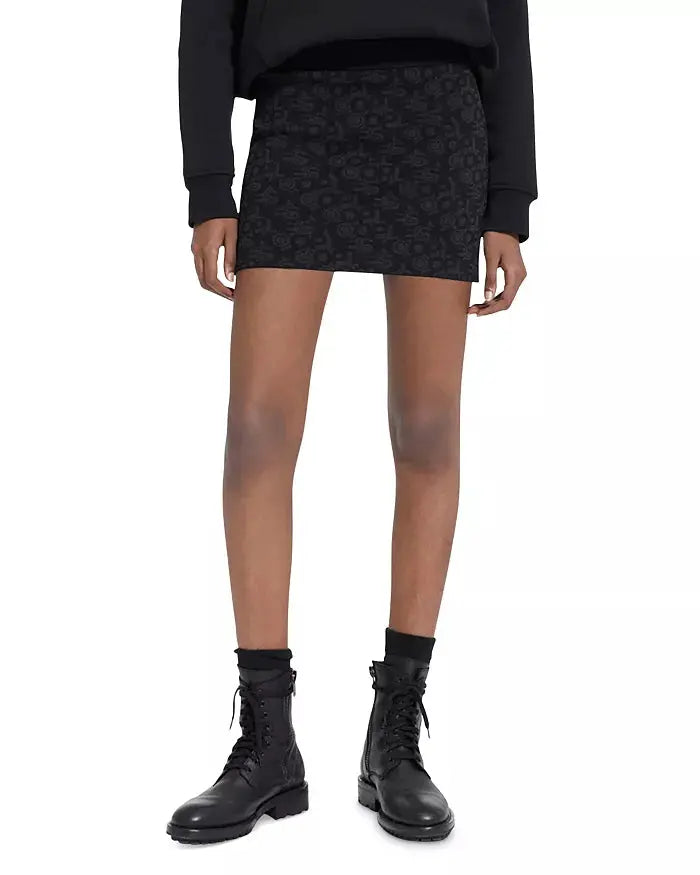 Mini-jupe noire à monogramme logo The Kooples, avec motifs floraux discrets et bottes lacées noires.