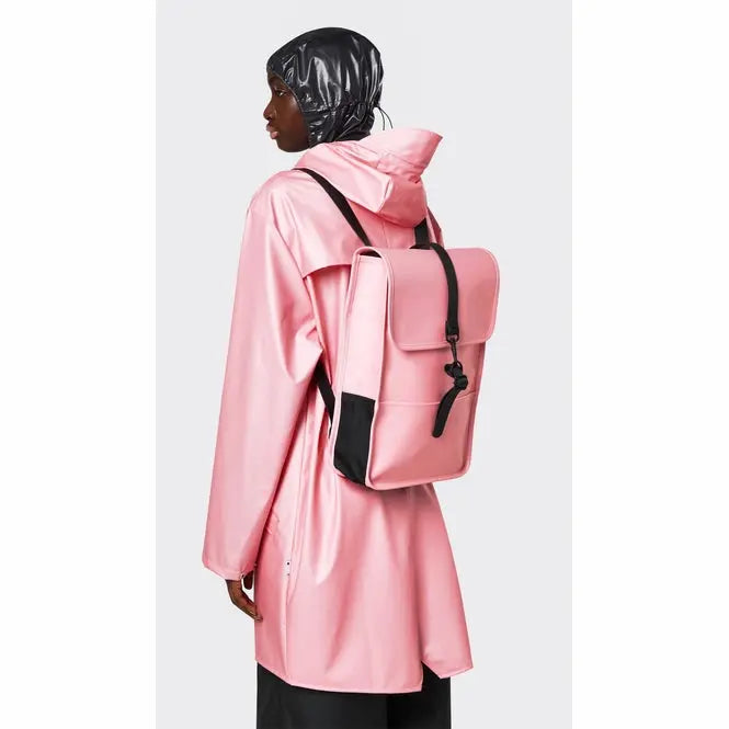 Mini sac à dos rose Rains avec bretelles noires, porté avec un imperméable rose et un bonnet noir brillant.