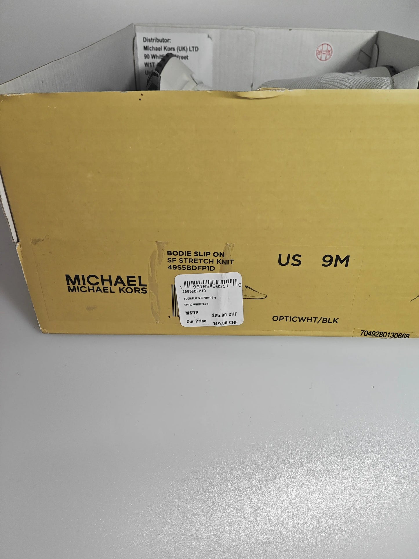 Chaussure slip-on en maille stretch blanc optique et noir de Michael Kors, visible dans sa boîte jaune.