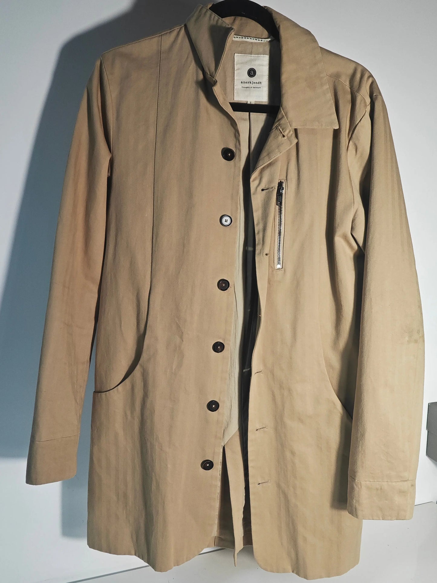 Manteau trench beige d'Anerkjendt, avec fermeture boutonnée et poche zippée visible, suspendu sur un cintre noir.