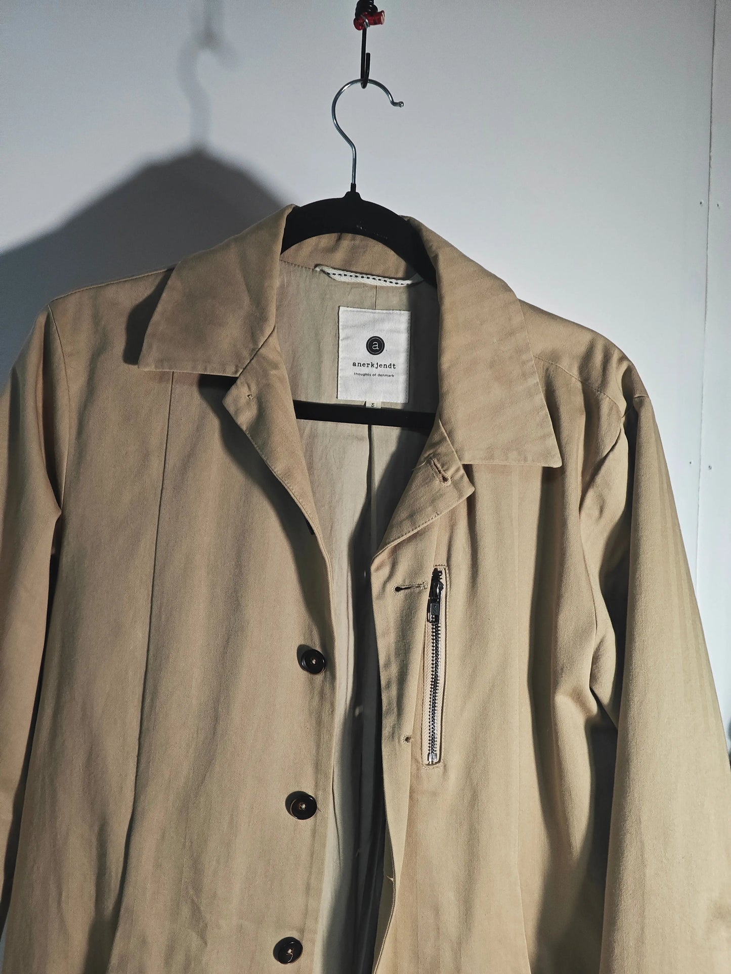 Manteau trench beige par Anerkjendt, avec une poche zippée et une fermeture à boutons marron.