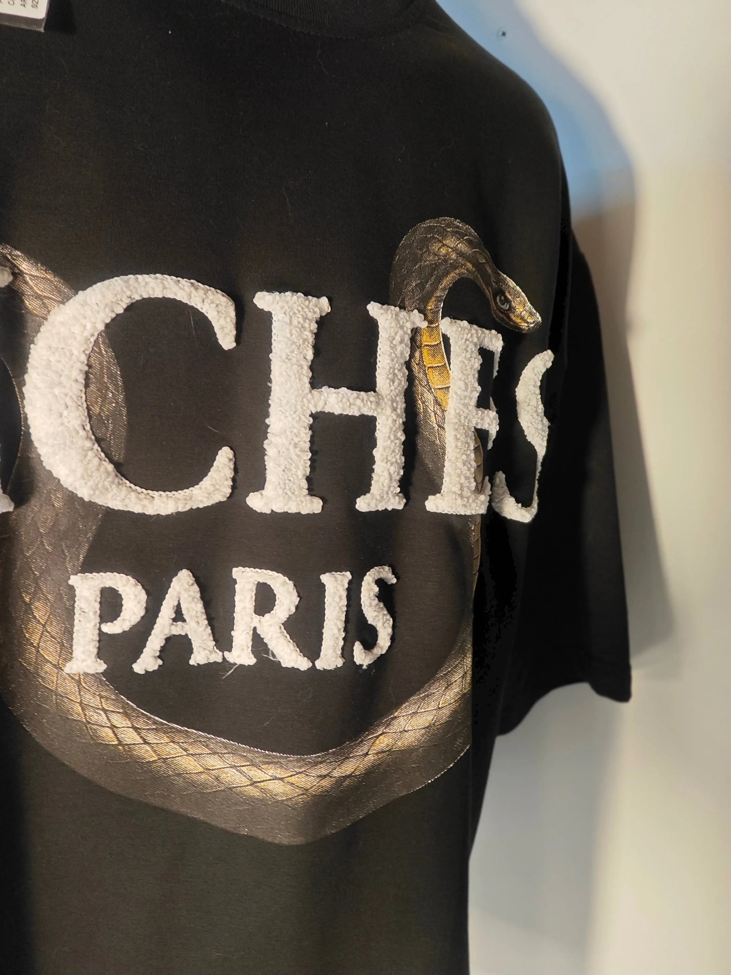 T-shirt noir de Riches Paris, avec un motif serpent imprimé et les mots « Riches Paris » en lettres blanches texturées.