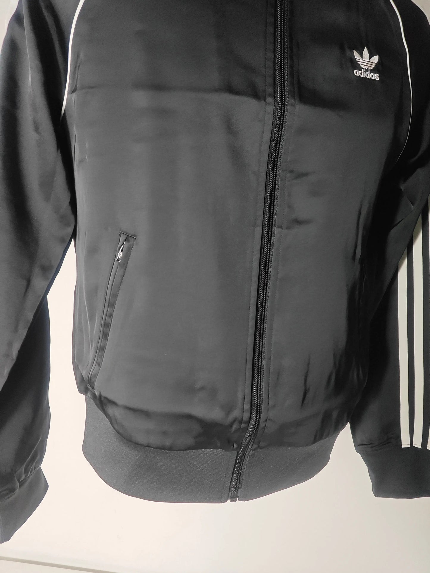 Veste de survêtement noire satinée, par Adidas, avec fermeture éclair et bandes blanches distinctives sur les manches.