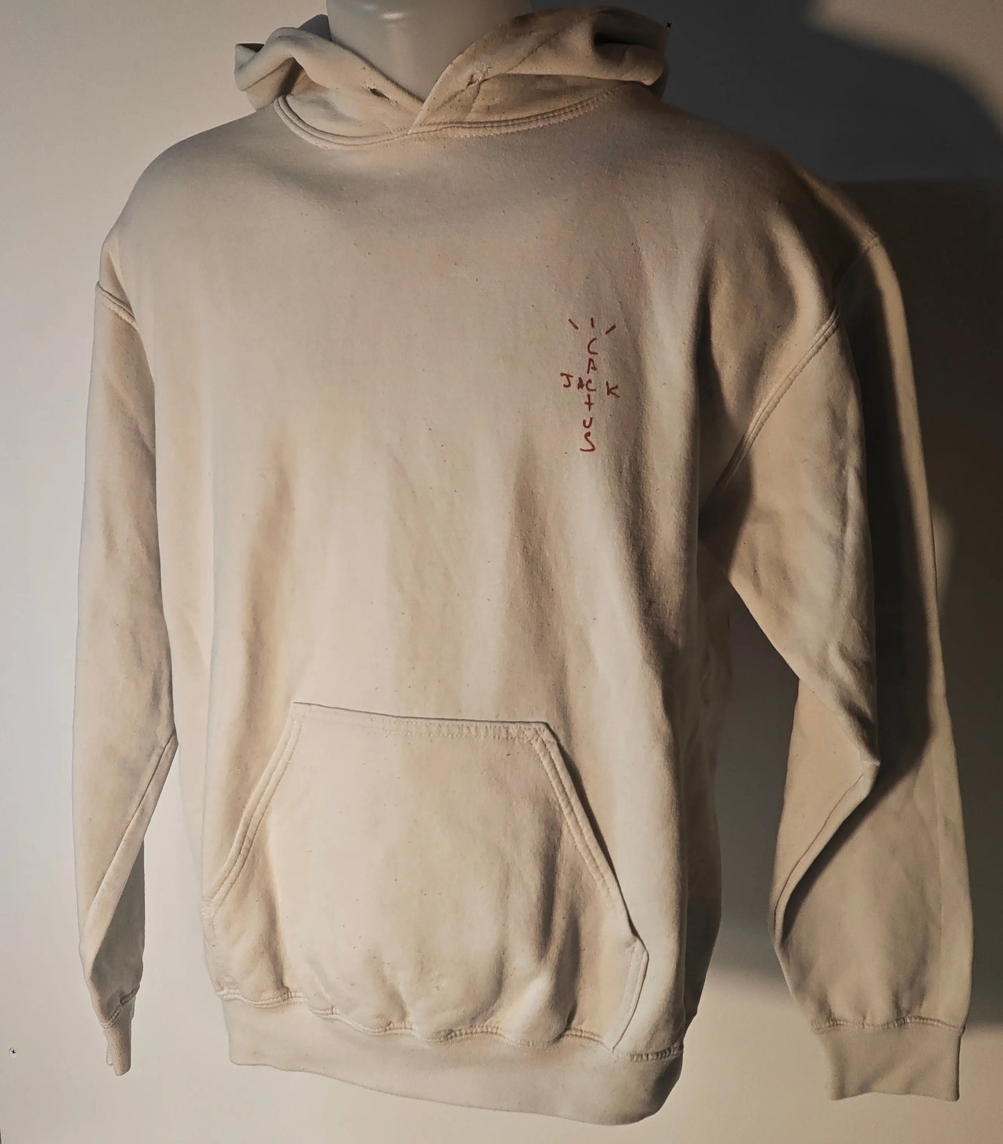 Sweat à capuche beige par Travis Scott, avec un logo rouge «Cactus Jack» sur la poitrine et une poche kangourou.