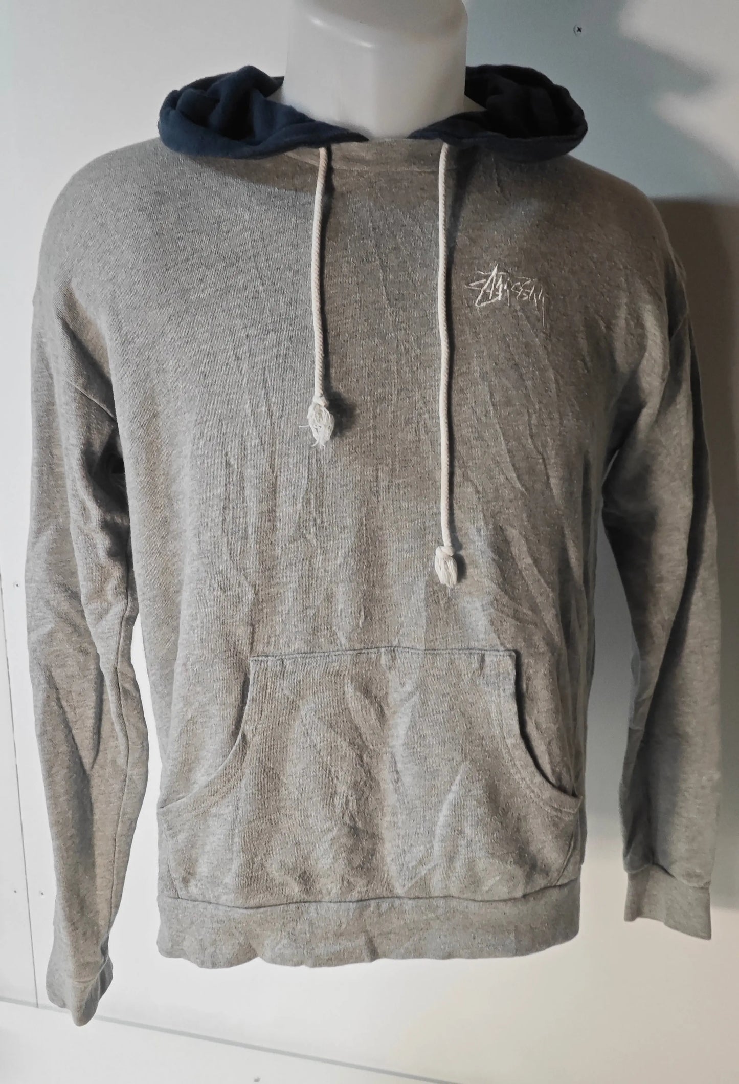 Sweat à capuche gris chiné de Stüssy, avec capuche bleu marine et cordon de serrage blanc, poche kangourou devant.