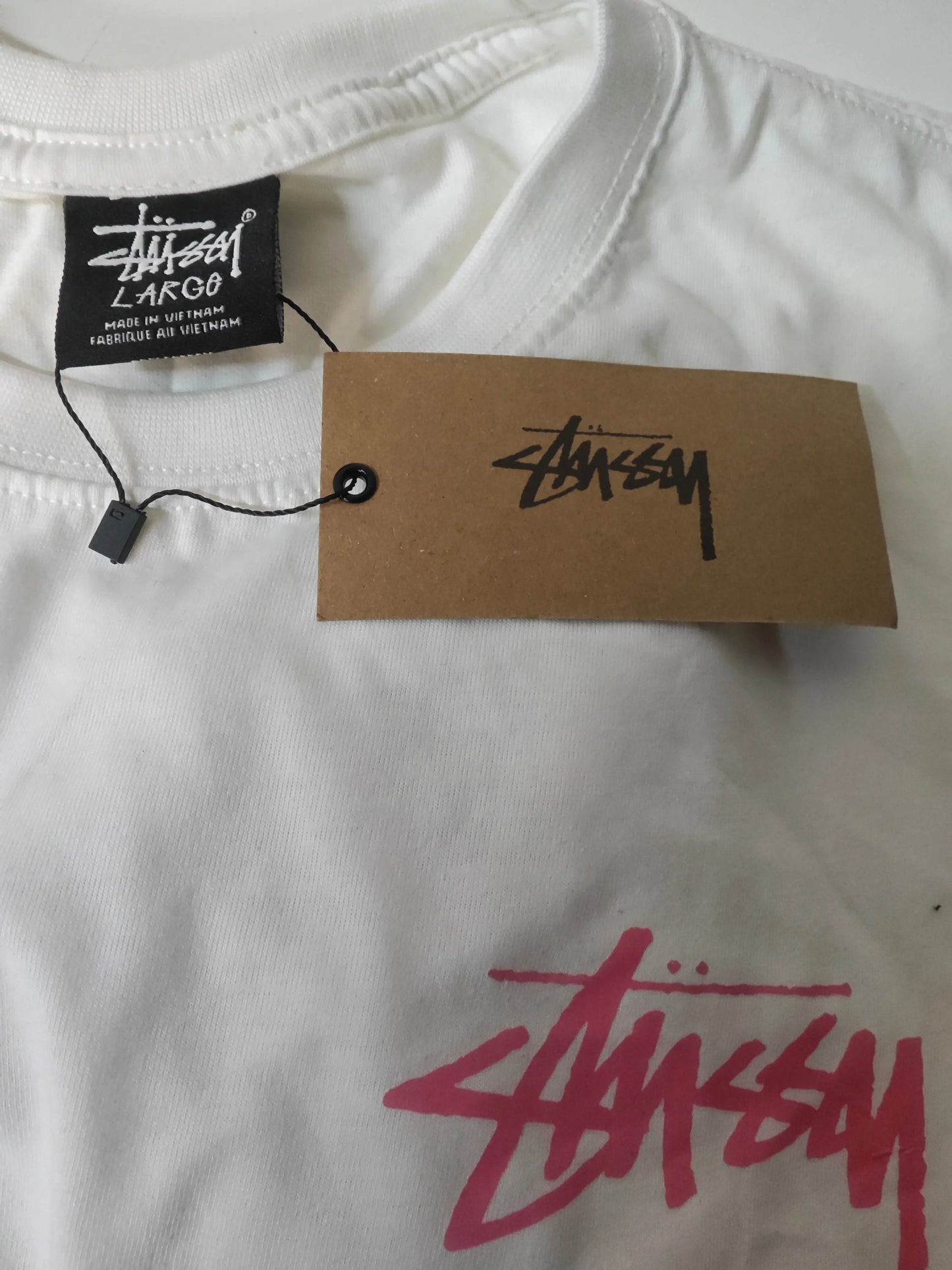 T-shirt blanc taille L de Stüssy avec étiquette noire et logo rose vif imprimé.