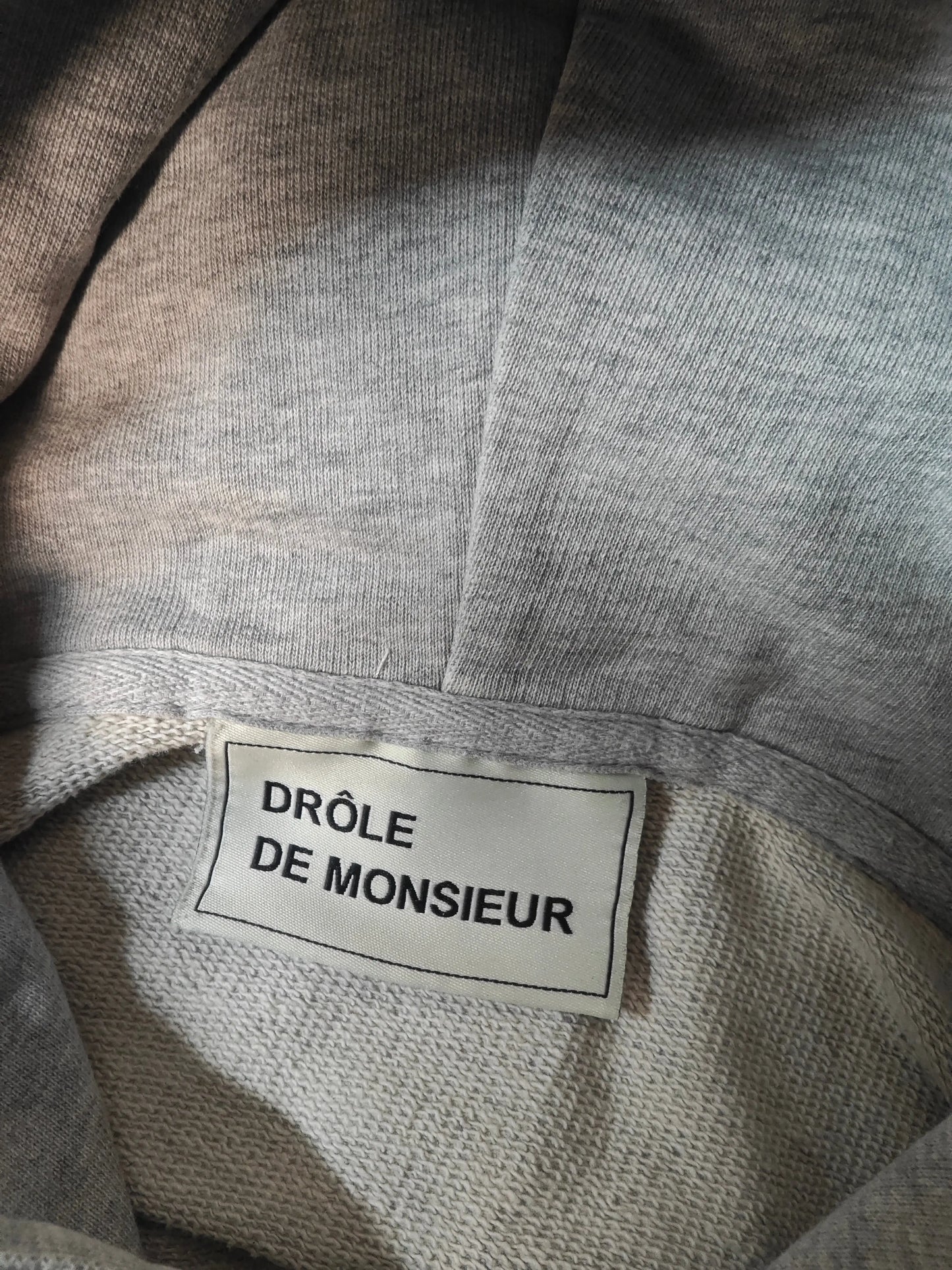 Sweat à capuche gris chiné de Drôle de Monsieur, avec une étiquette en tissu à bordure noire.