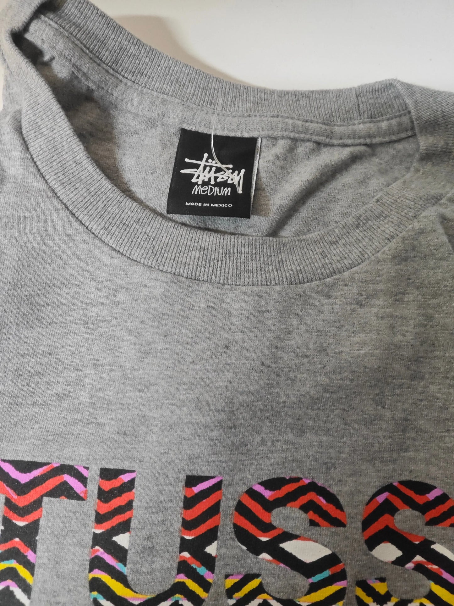 T-shirt gris chiné taille M de Stüssy, avec inscription multicolore en bas.