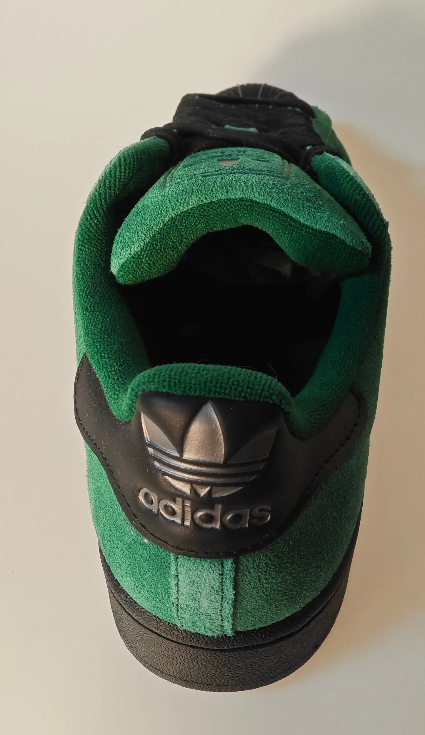 Chaussure de sport en suède vert et noir, de Adidas, avec logo trèfle argenté sur l'arrière et semelle texturée noire.