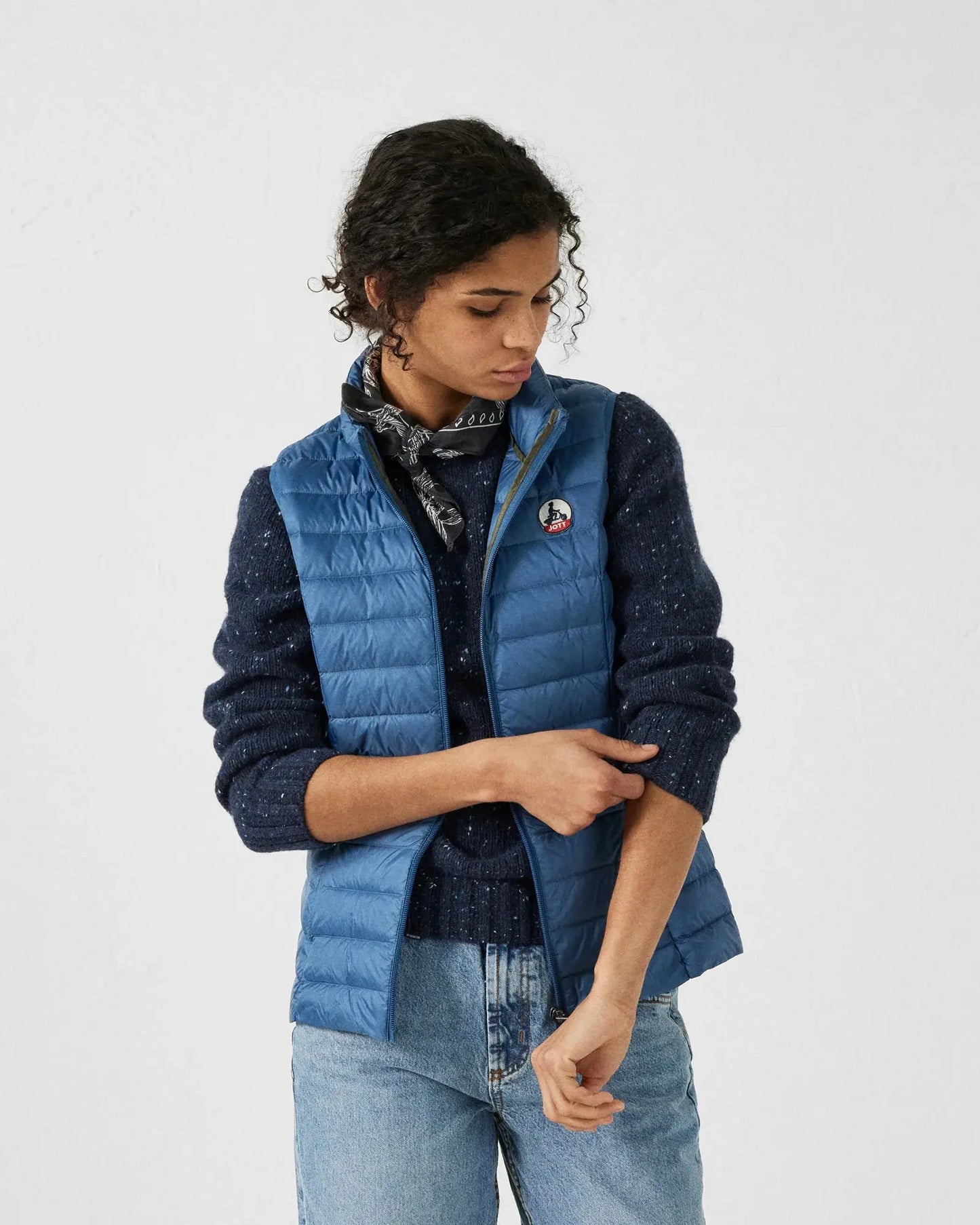 Doudoune sans manches bleue jeans by Jott, portée avec un pull moucheté et un foulard à motifs.