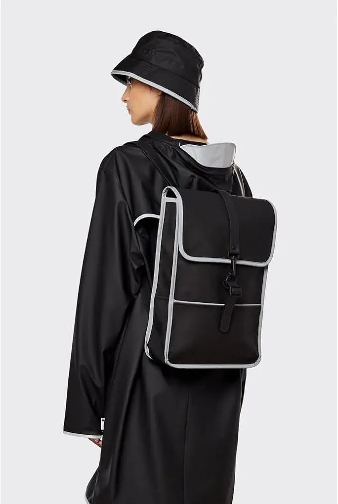Sac à dos noir réfléchissant de Rains, porté avec un imperméable noir et un chapeau assorti.