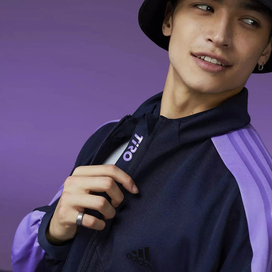 Veste de survêtement bleu marine avec rayures violettes par Adidas, portée avec un chapeau noir et une bague argentée.