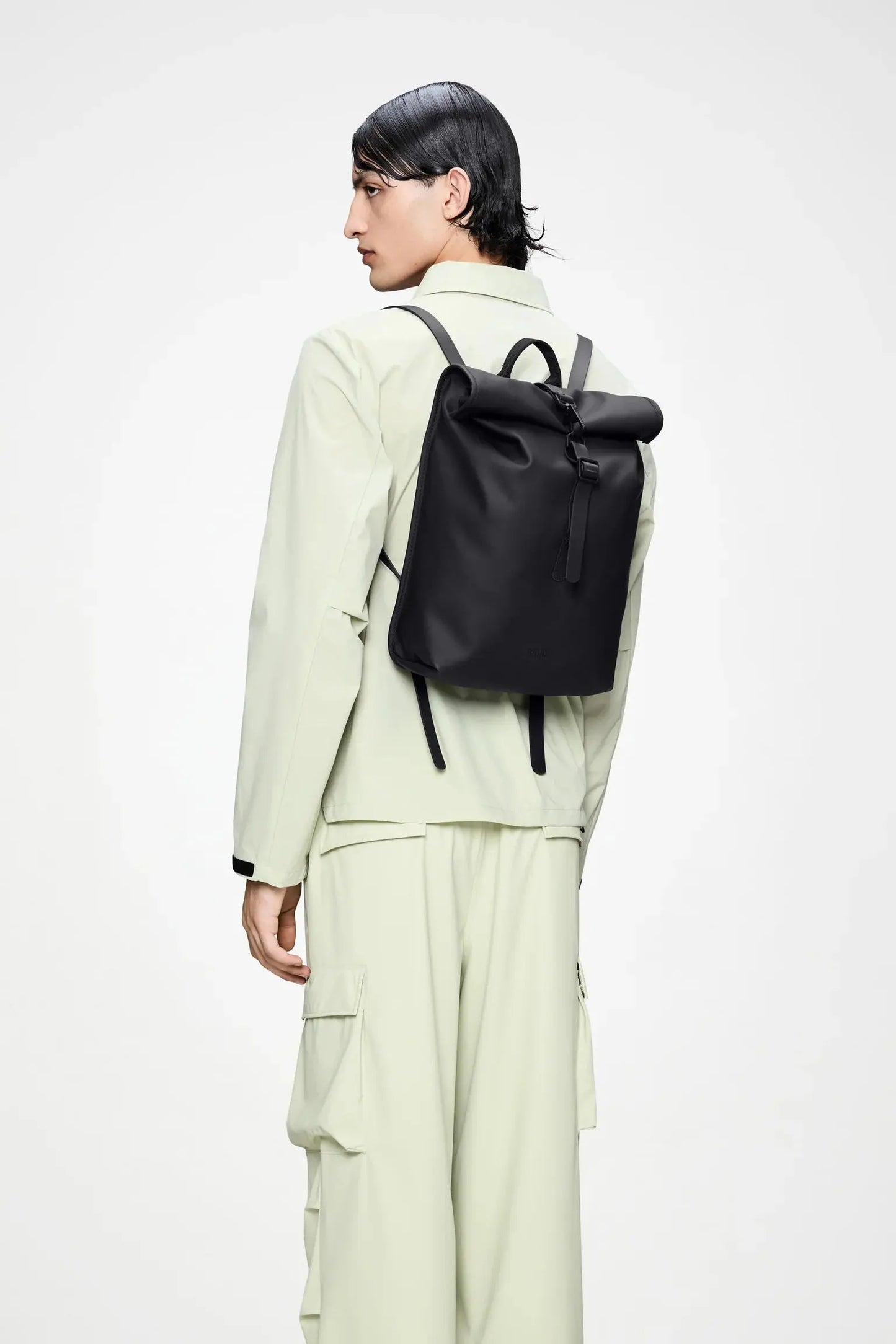 Mini sac à dos rolltop noir de Rains, porté sur le dos avec une veste et un pantalon vert clair.