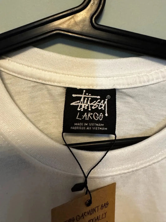 T-shirt blanc par Stüssy, étiquette noire de la marque visible, suspendu sur un cintre noir.