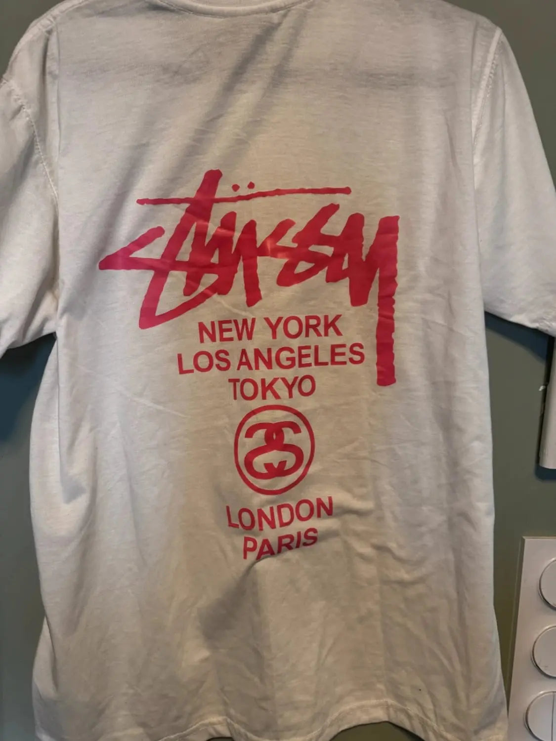 T-shirt blanc imprimé de Stüssy, avec logo et noms de villes en rose vif.