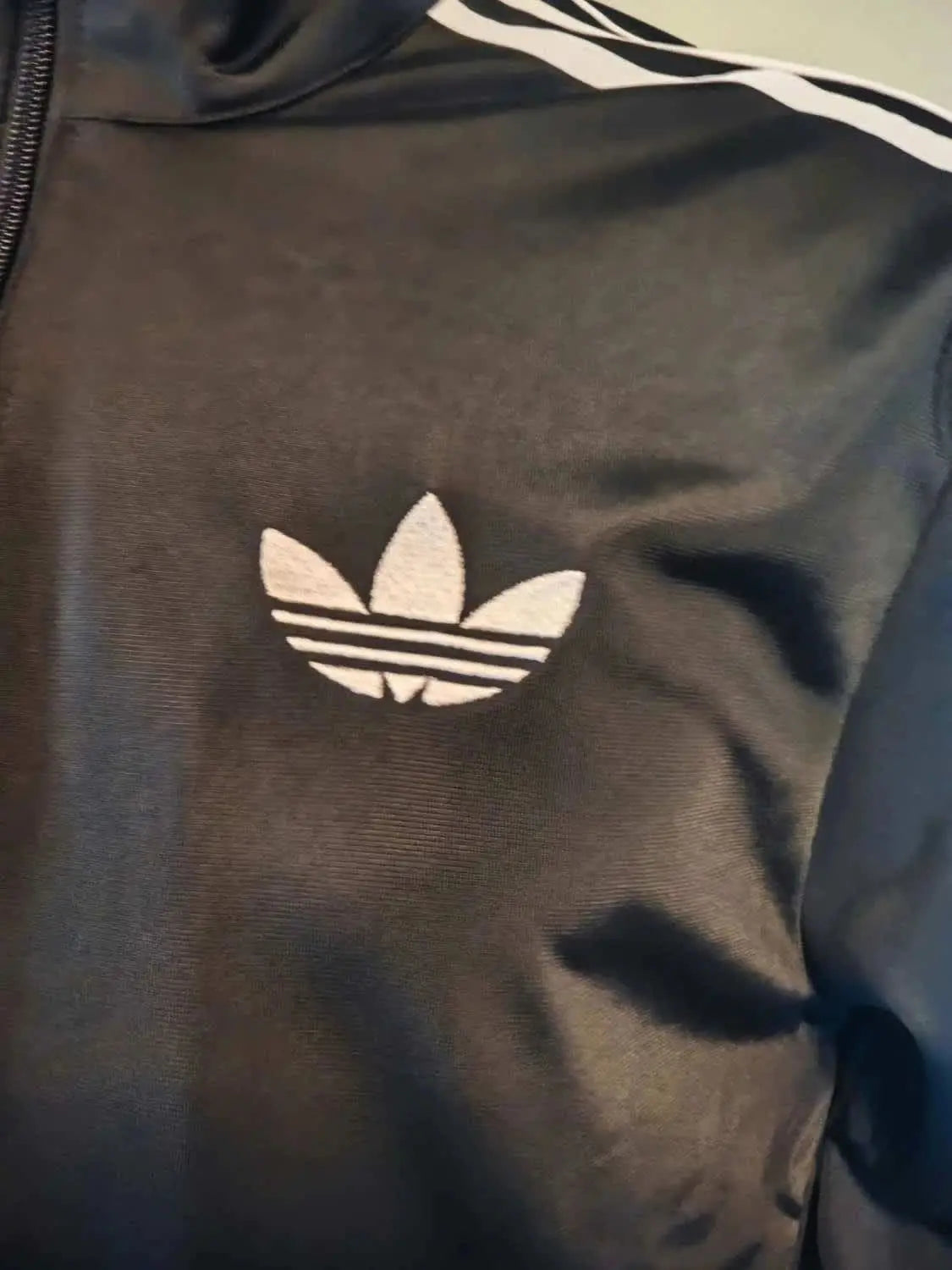 Veste de survêtement noire de Adidas Originals avec un logo blanc et trois bandes blanches sur l'épaule.
