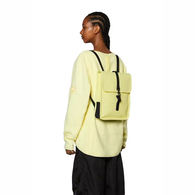 Mini sac à dos jaune pastel de Rains avec des bretelles noires, porté avec un survêtement jaune.