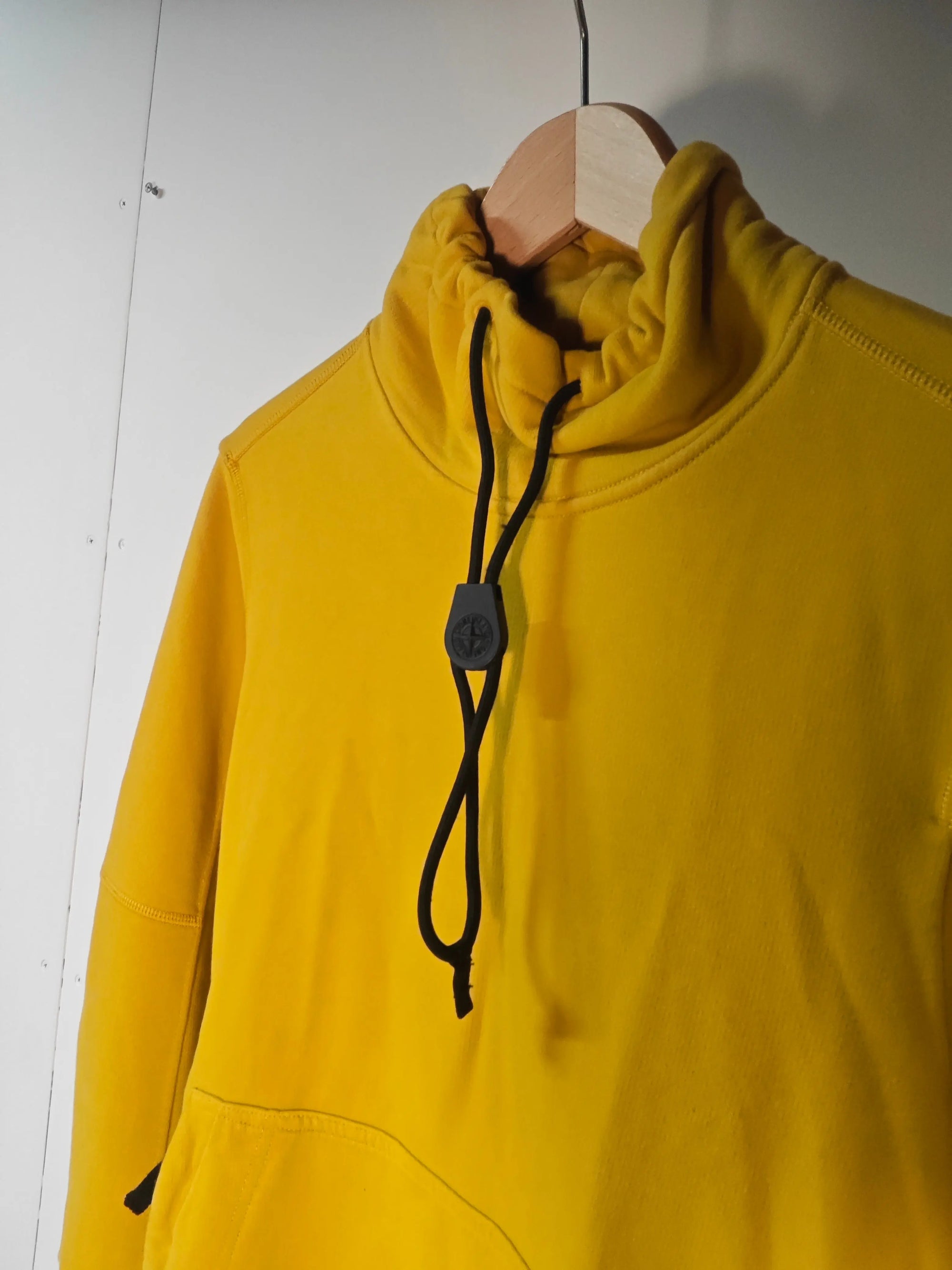 Sweat jaune de Stone Island, suspendu sur un cintre en bois, avec cordon noir au niveau du col.
