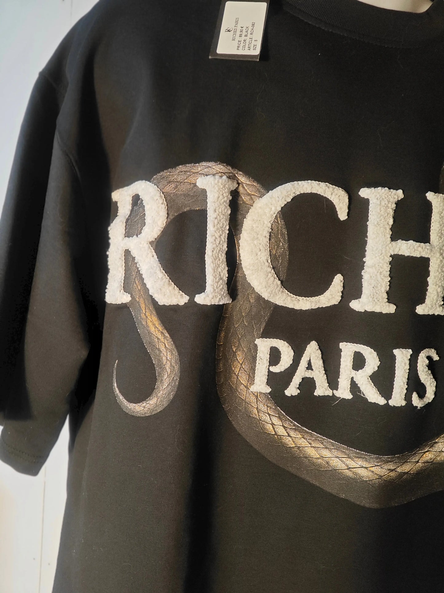 T-shirt noir pour homme de Riches Paris avec inscription en lettres blanches texturées et motif serpent doré.
