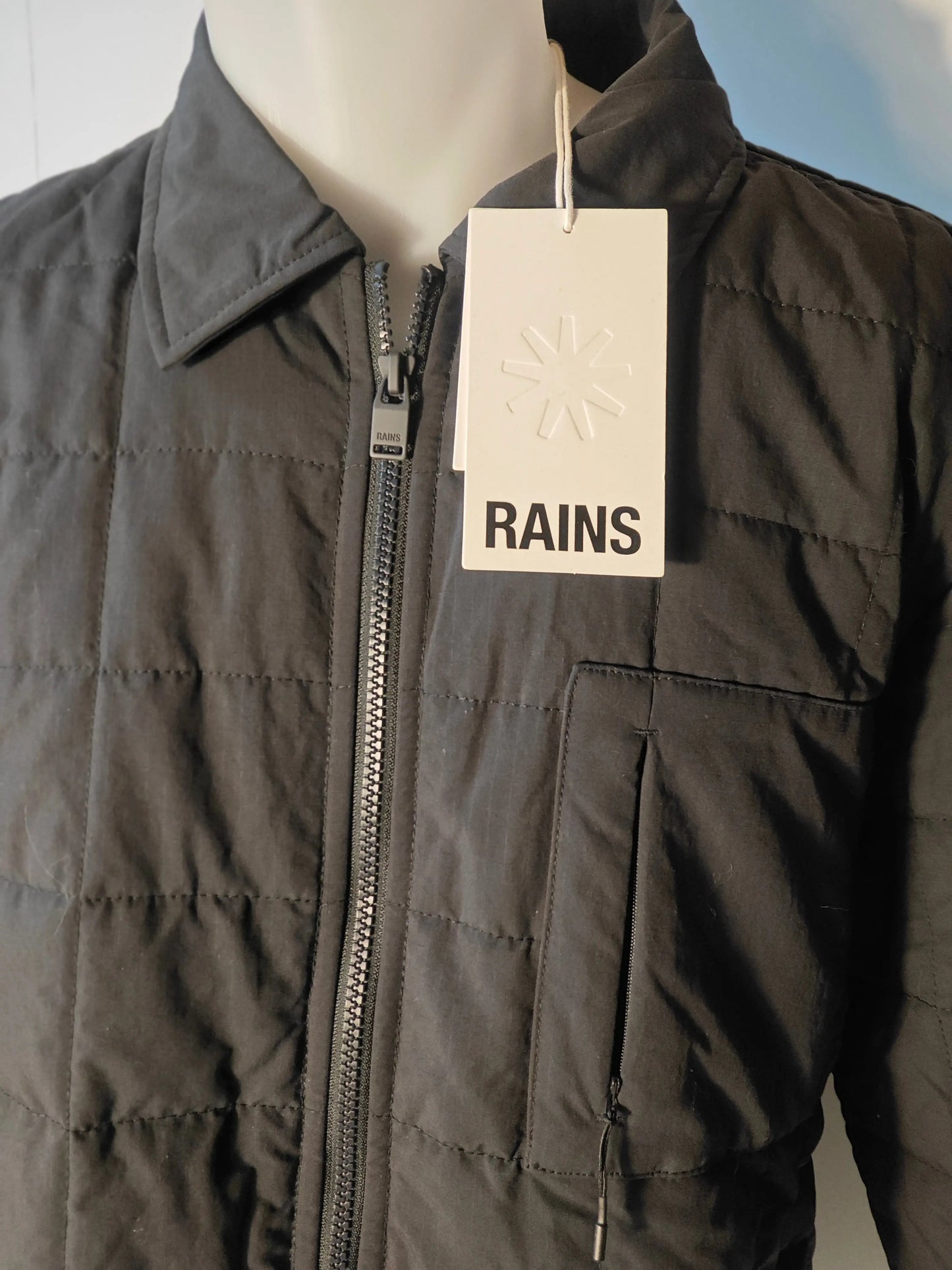 Veste matelassée bleu marine de Rains, avec fermeture éclair sur le devant et étiquette blanche.