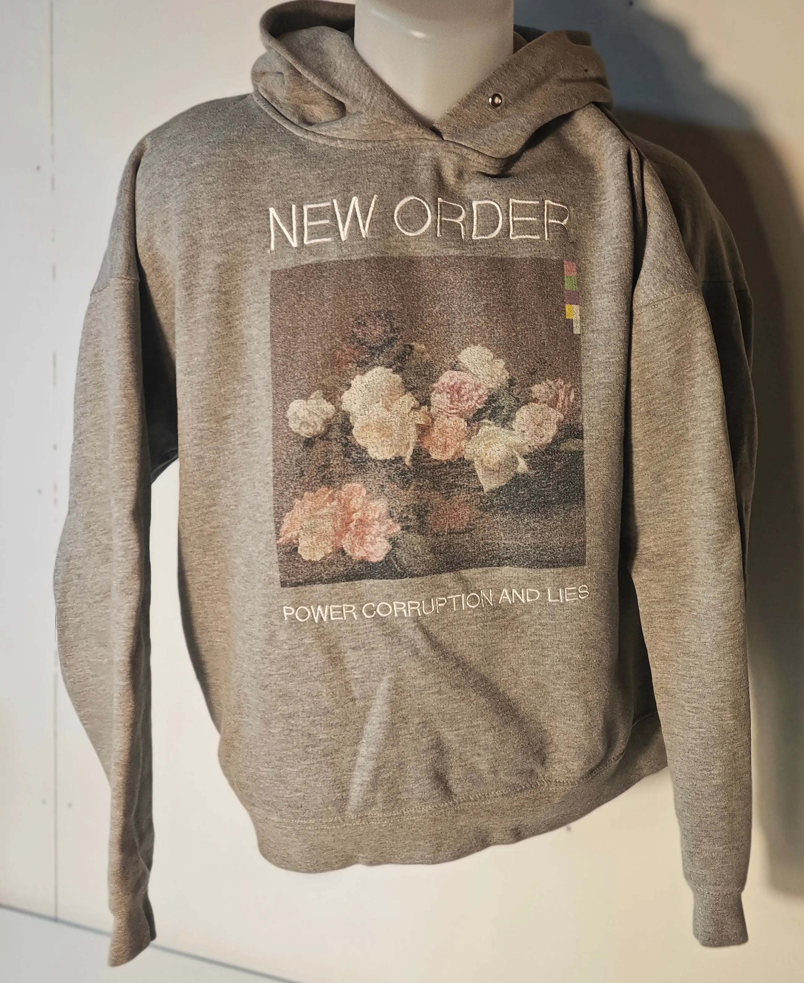 Sweat à capuche gris de Sandro x New Order, avec motif floral et lettrage « New Order » et « Power Corruption and Lies ».
