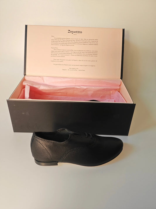 Richelieu en cuir noir Repetto, sortant de sa boîte noire avec papier de soie rose.
