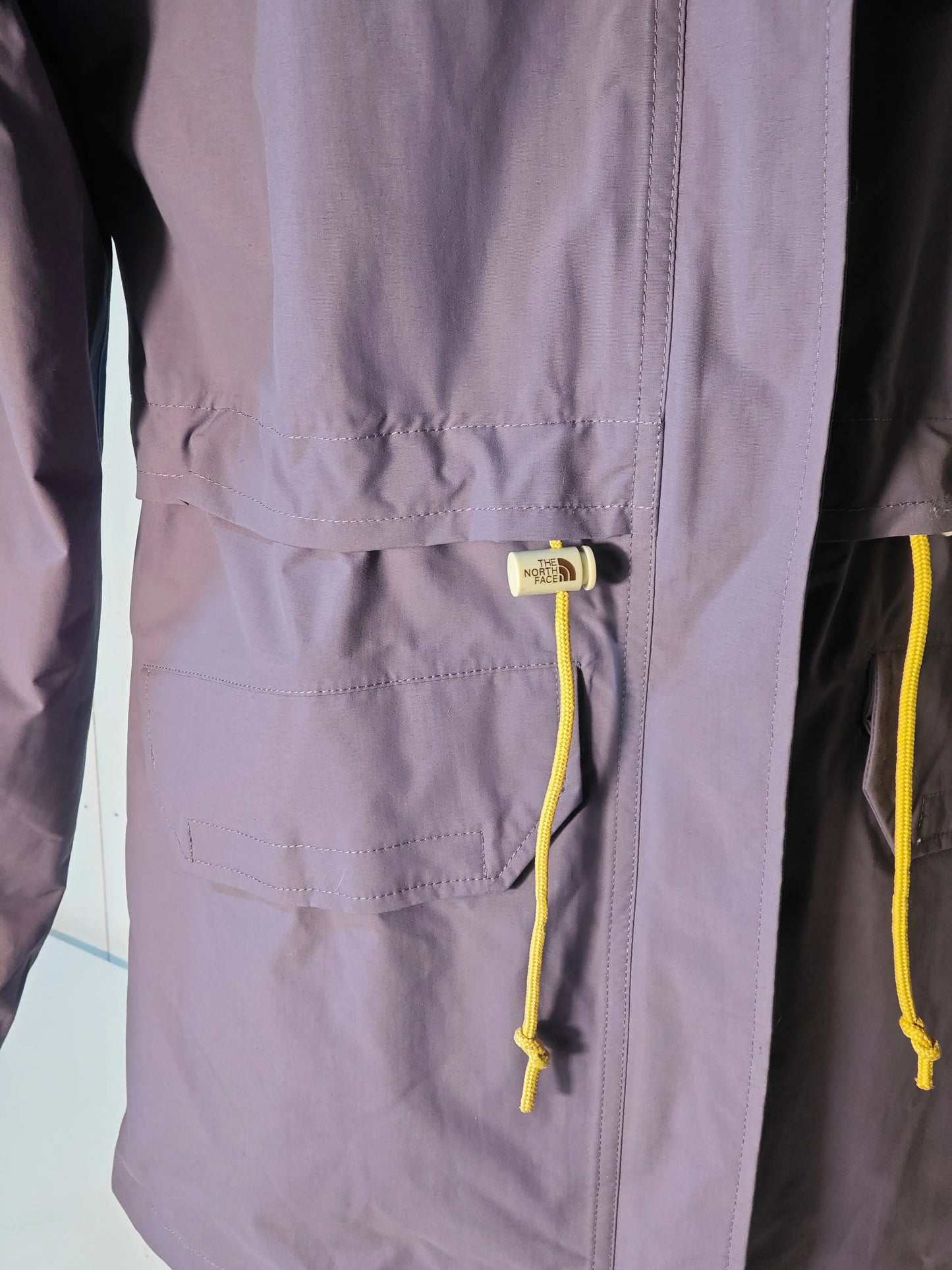 Parka DryVent violet lavande pour femme de The North Face, avec cordon de serrage jaune et poche frontale.
