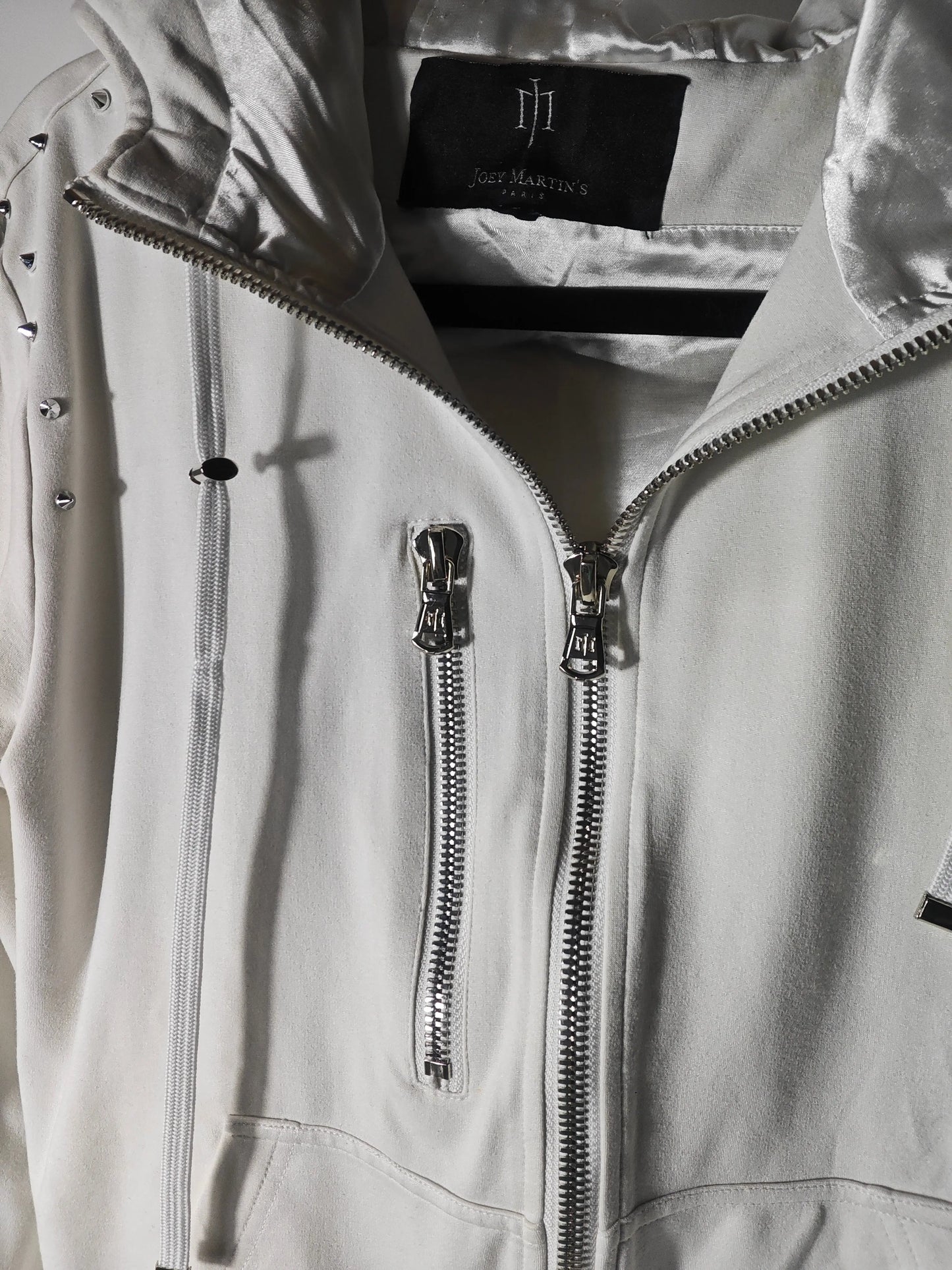 Veste à capuche blanche de Joey Martin’s Paris avec cordons de serrage et fermetures éclair argentées, ornée de clous.
