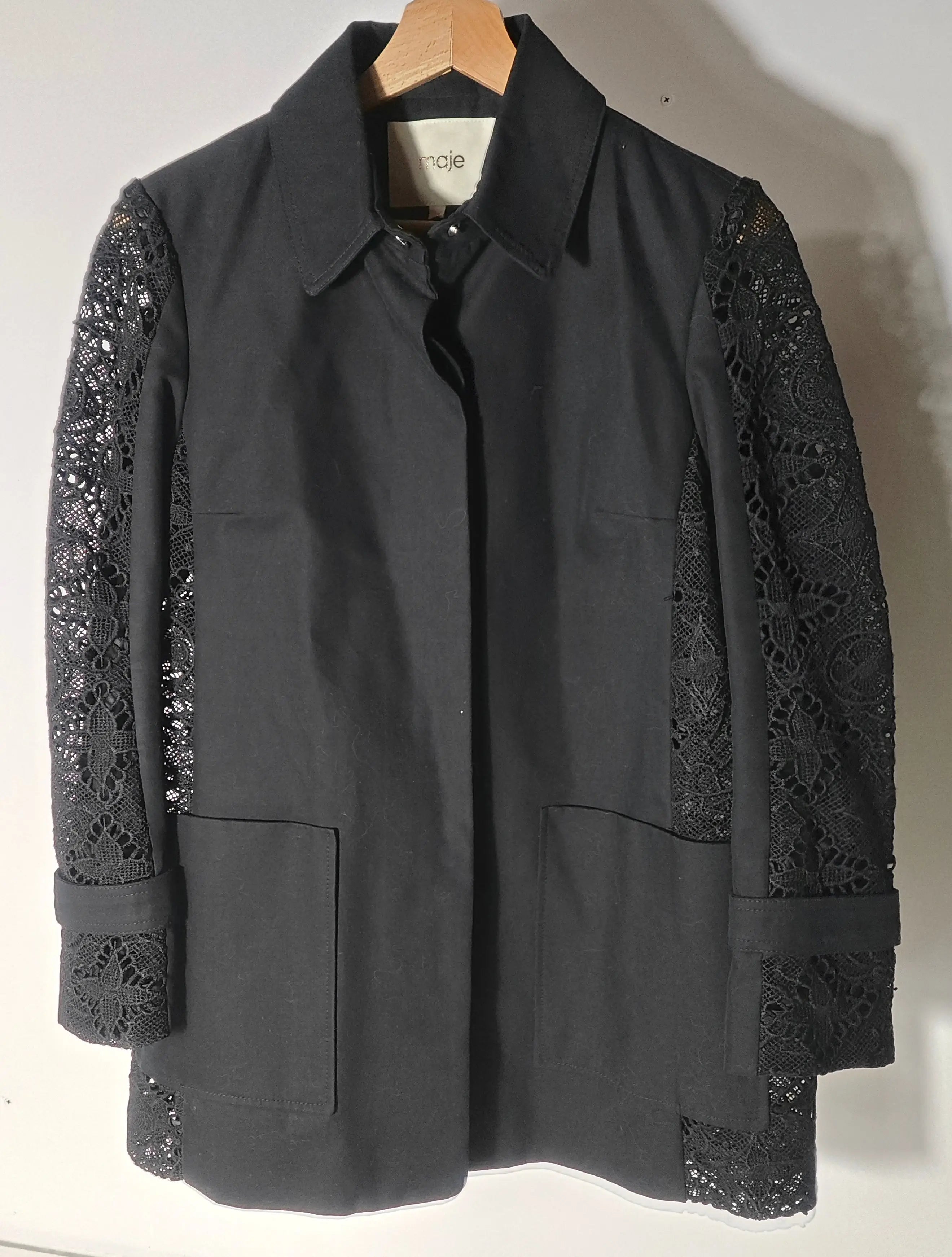 Veste noire à manches en dentelle par Maje, sur un cintre en bois, avec des poches carrées.
