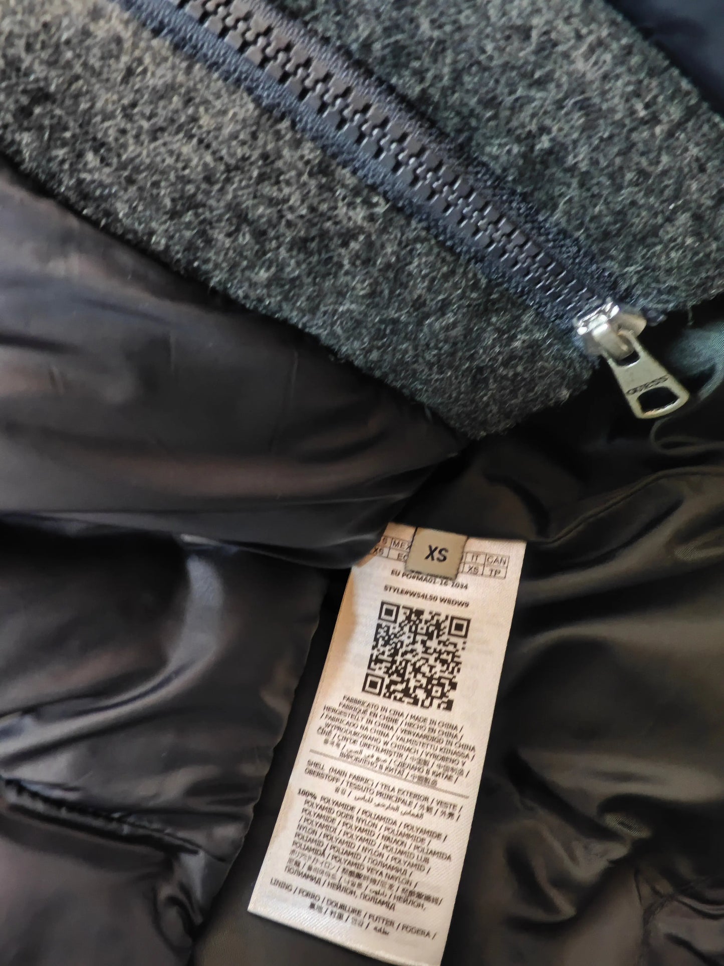 Doudoune longue noire avec détails en laine de Guess, présentant une étiquette de taille et un code QR.
