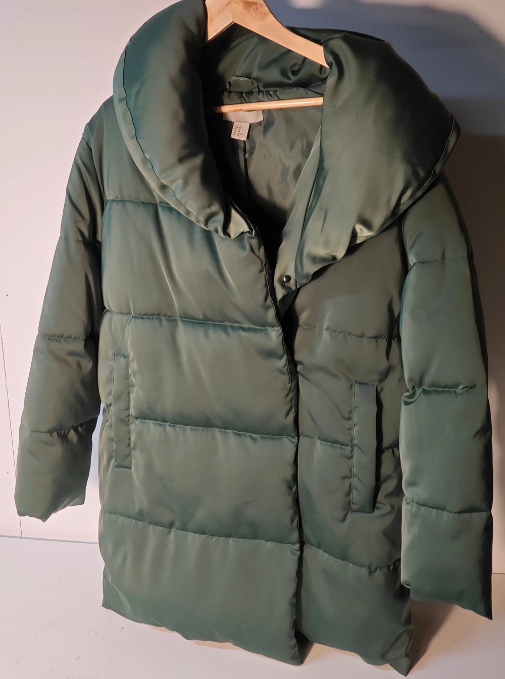 Doudoune verte de H&M sur un cintre en bois, avec un col volumineux et une coupe longue.
