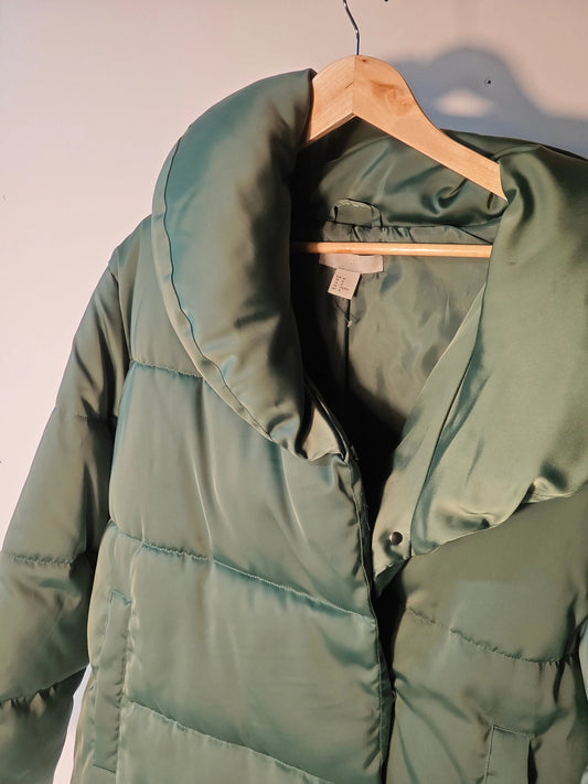 Doudoune verte de H&M sur un cintre en bois, avec un large col et une fermeture boutonnée.
