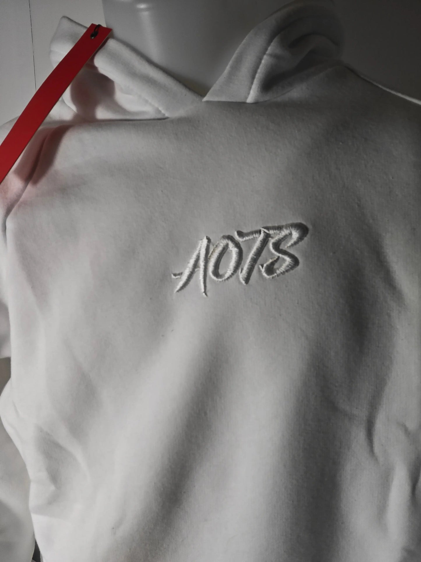 Sweat à capuche blanc avec broderie, de AOTS, sur un mannequin, avec une lanière rouge visible.