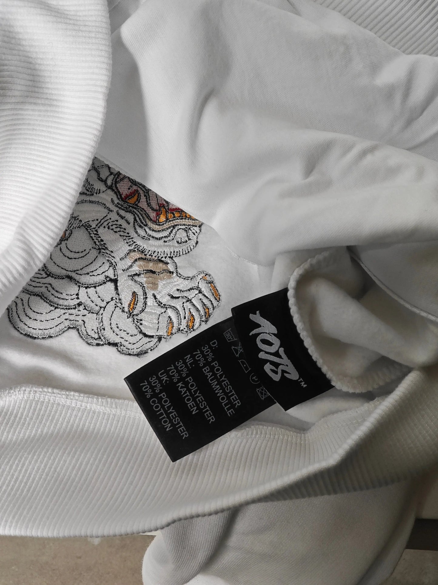 Sweat à capuche blanc de AOTS avec une broderie japonaise détaillée et une étiquette d'entretien visible.