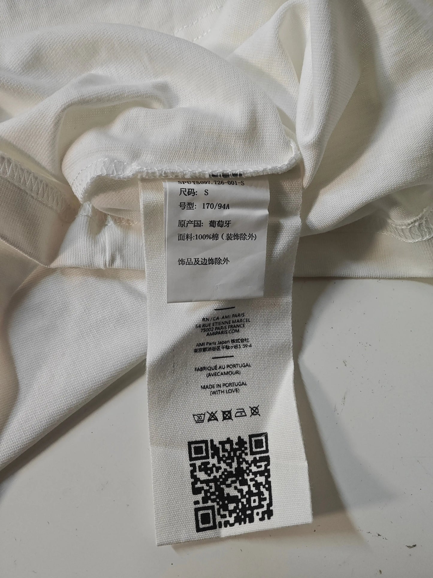 T-shirt blanc en coton, d'AMI, avec étiquette indiquant les instructions de lavage et un QR code.
