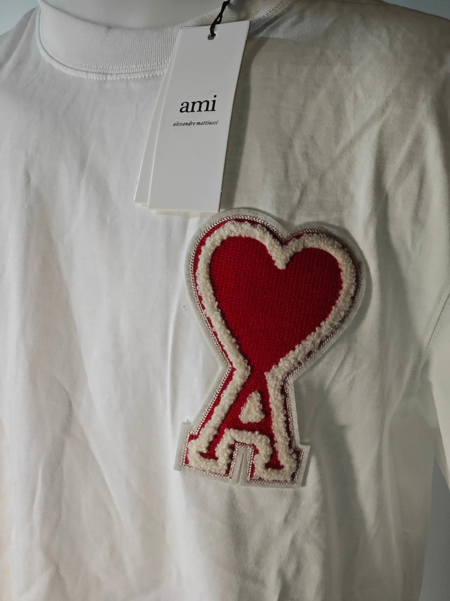 T-shirt blanc de AMI Paris, avec un patch bouclette rouge en forme de cœur et un tag de la marque.
