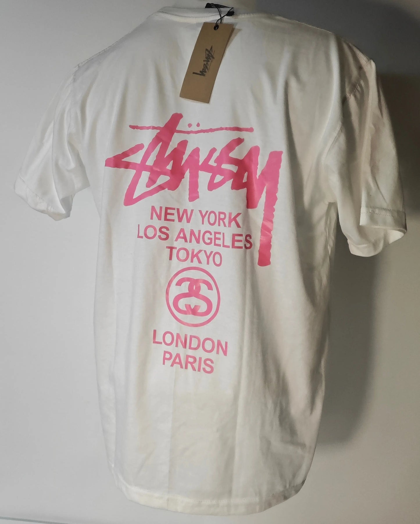 T-shirt blanc à manches courtes de Stüssy, avec imprimé rose du logo et villes au dos.
