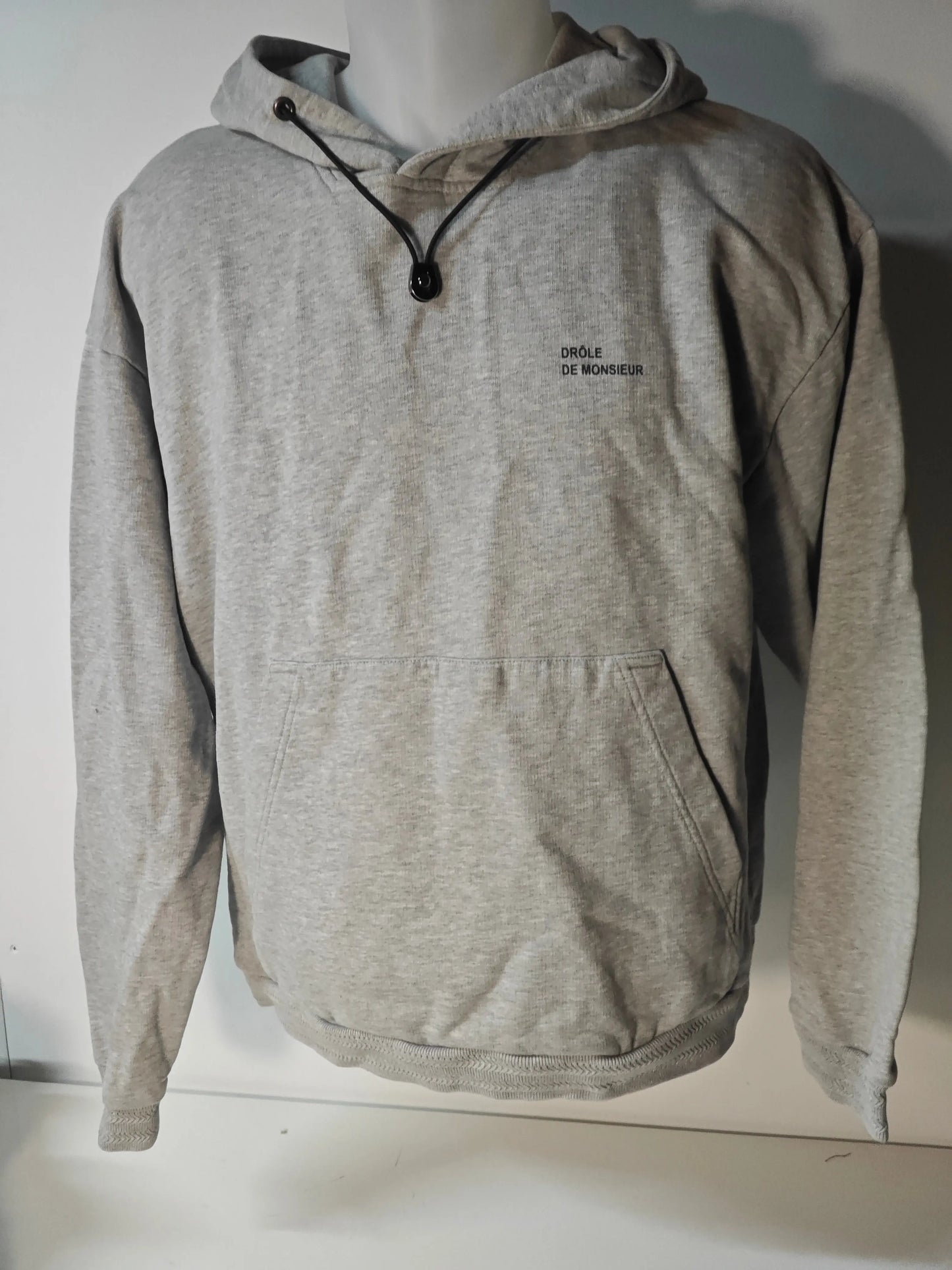 Sweat à capuche gris chiné de Drôle de Monsieur, avec cordon de serrage noir et poche kangourou.
