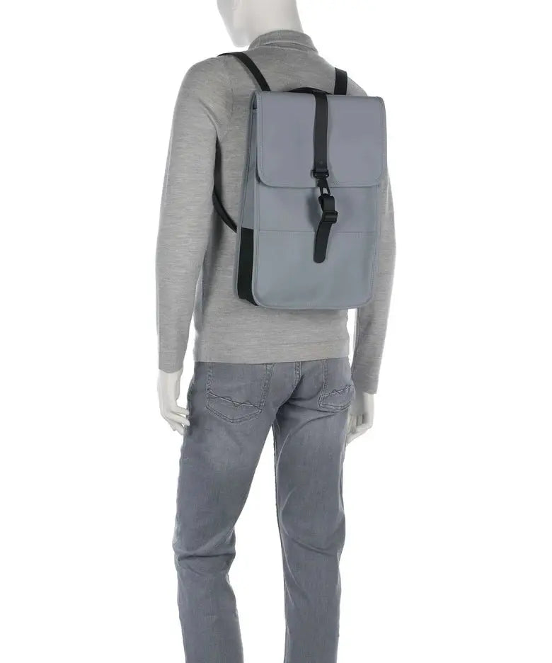 Sac à dos gris Rains Backpack Mini River par Rains, avec bretelles noires, porté avec un pull et un jean gris.