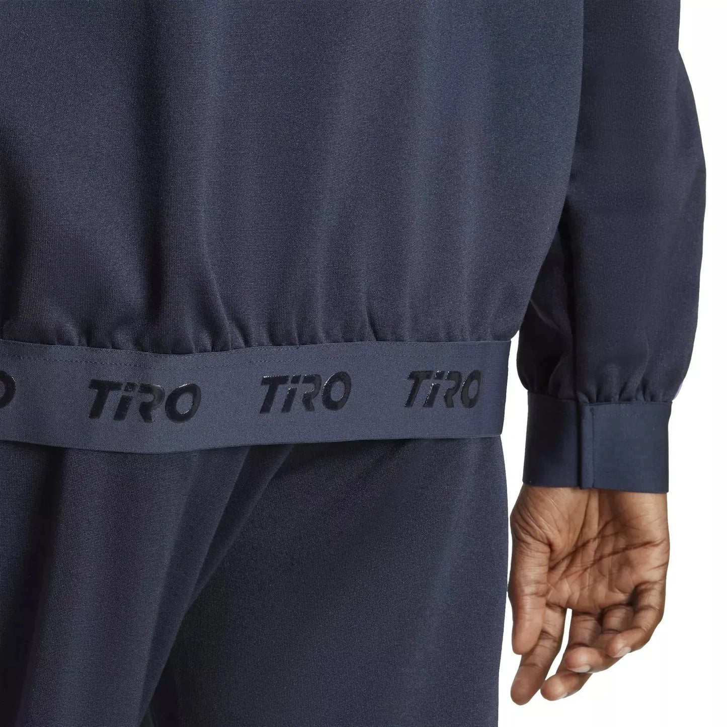 Veste de survêtement bleue de Adidas, avec une bande élastique portant le logo "TIRO" répété, et une manche longue.