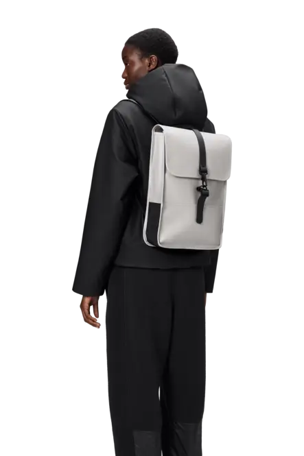 Sac à dos gris Rains BackPack Mini, par Rains, porté avec une veste et un pantalon noirs.