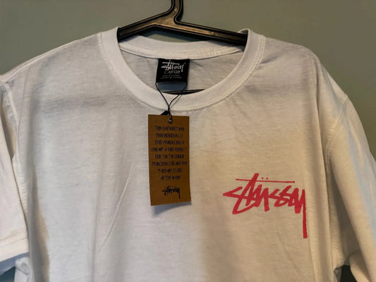 T-shirt blanc de Stüssy avec un logo rose vif sur le devant et une étiquette cartonnée.
